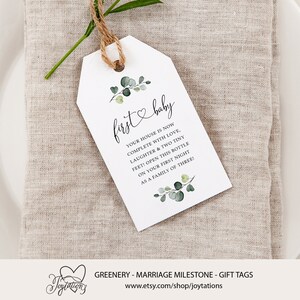 May include: A white gift tag with green eucalyptus leaves and a heart with the text "first baby" and the message "Your house is now complete with love, laughter & two tiny feet! Open this bottle on your first night as a family of three!"