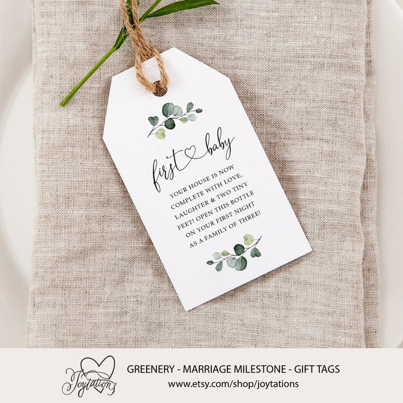 May include: A white gift tag with green eucalyptus leaves and a heart with the text "first baby" and the message "Your house is now complete with love, laughter & two tiny feet! Open this bottle on your first night as a family of three!"