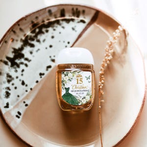 May include: A small, clear hand sanitizer bottle with a white cap, featuring a label with the text "15 Christina Mi Quinceanera 10.12.25". The label includes a green dress illustration and floral accents. The bottle sits on a brown and white speckled plate.