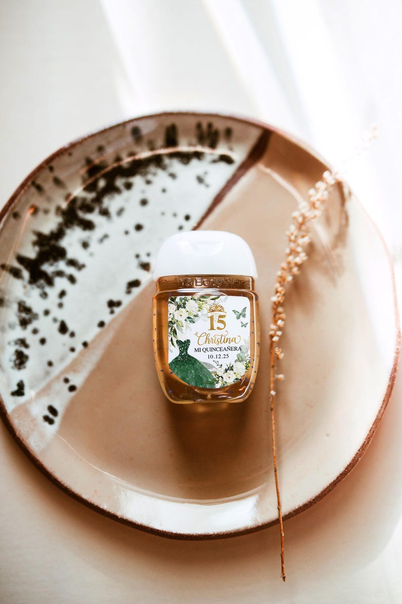 May include: A small, clear hand sanitizer bottle with a white cap, featuring a label with the text "15 Christina Mi Quinceanera 10.12.25". The label includes a green dress illustration and floral accents. The bottle sits on a brown and white speckled plate.