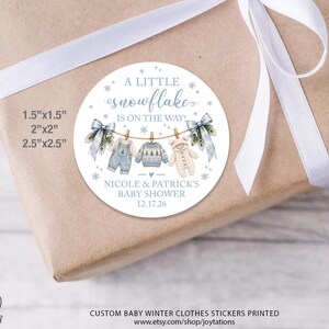 May include: A round white sticker featuring blue text that reads A LITTLE SNOWFLAKE IS ON THE WAY, NICOLE & PATRICK'S BABY SHOWER, and 12.17.26. The design includes illustrations of baby clothes on a clothesline and snowflakes.