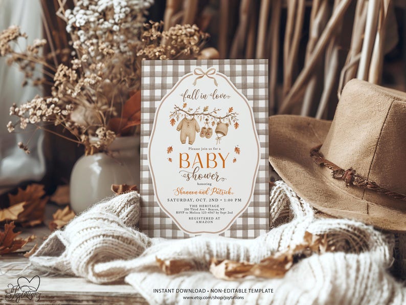 May include: A rectangular baby shower invitation card with a brown and white gingham border. The center features a white label shape with a bow, autumn leaves, and baby clothes illustrations. Text includes fall in love, baby shower, Shannon and Patrick, Saturday, Oct. 2nd, 1:00 PM, The Heritage, 100 Third Ave, Beacon, NY, RSVP to Melissa 123-4567 by Sept 2nd, and Registered at Amazon.
