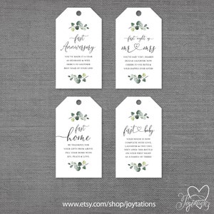May include: Four white gift tags with green eucalyptus leaves and calligraphy text. The tags read "first anniversary", "first night as mr & mrs", "first home", and "first baby".