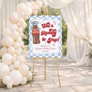 Puede incluir: Un letrero de bienvenida para baby shower sobre un caballete dorado. El letrero tiene un borde a cuadros azul y blanco y presenta una ilustraci&oacute;n de botella de cola y el texto "She's Ready to Pop!" con los detalles del baby shower.