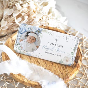 May include: A personalized candy bar wrapper with a baby's photo, blue floral accents, and the text "GOD BLESS Miguel Rivera September 6, 2025." The wrapper is on a wooden tray with white ribbon and dried flowers.