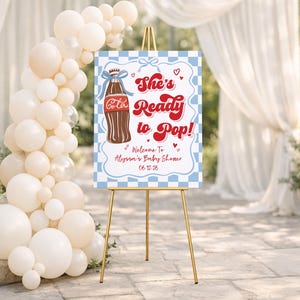 Puede incluir: Un letrero de bienvenida para un baby shower sobre un caballete dorado. El letrero tiene un borde a cuadros azul y blanco y presenta una ilustraci&oacute;n de una botella de cola y el texto "She's Ready to Pop!" con los detalles del baby shower.