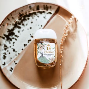 May include: A small, clear hand sanitizer bottle with a white cap, featuring a label with the text "15 Christina Mi Quincea&ntilde;era 10.12.25". The label includes a floral design and a drawing of a dress. The bottle sits on a brown plate.