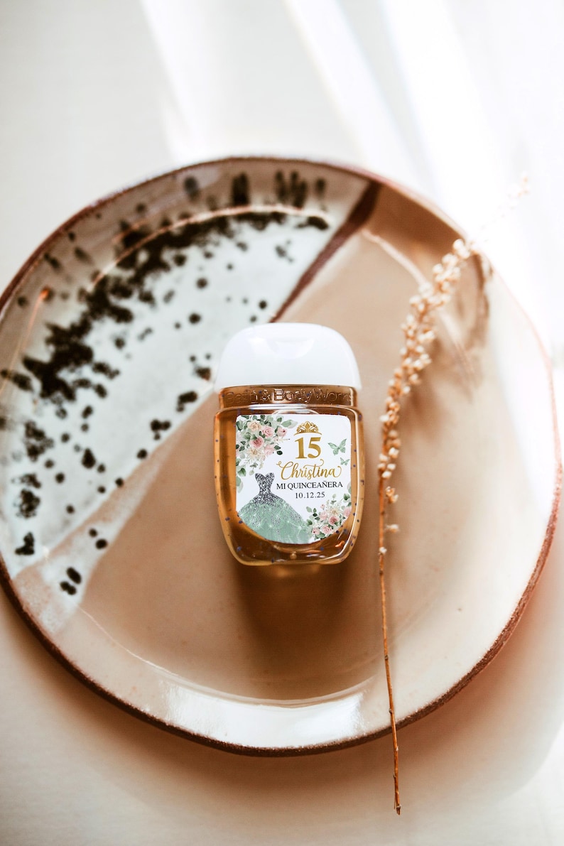 May include: A small, clear hand sanitizer bottle with a white cap, featuring a label with the text "15 Christina Mi Quincea&ntilde;era 10.12.25". The label includes a floral design and a drawing of a dress. The bottle sits on a brown plate.