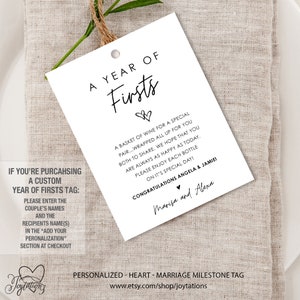 May include: A white gift tag with black text that reads "A Year of Firsts". The tag is decorated with two small hearts. The text also reads "A basket of wine for a special pair...wrapped all up for you both to share. We hope that you are always as happy as today. Please enjoy each bottle on it's special day! Congratulations Angela & Jamie! Marisa and Alexa". The tag is personalized with the couple's names and a message for the recipient.