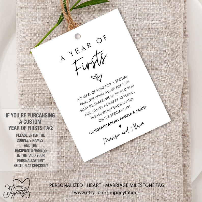 May include: A white gift tag with black text that reads "A Year of Firsts". The tag is decorated with two small hearts. The text also reads "A basket of wine for a special pair...wrapped all up for you both to share. We hope that you are always as happy as today. Please enjoy each bottle on it's special day! Congratulations Angela & Jamie! Marisa and Alexa". The tag is personalized with the couple's names and a message for the recipient.