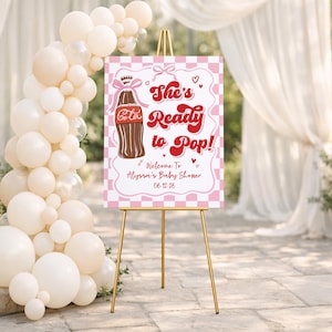 May include: A baby shower sign on a gold easel. The sign is pink and white with the text "She's Ready to Pop!" and a cartoon cola bottle. A balloon arch and draped fabric are in the background.