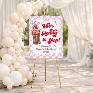 May include: A baby shower welcome sign on a gold easel. The sign is pink and white with a checkerboard pattern and features a brown soda bottle illustration and the text "She's Ready to Pop!" with the baby shower date.