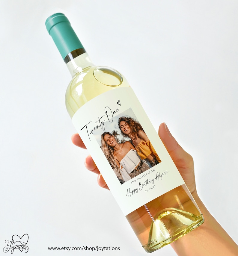 May include: White wine bottle with a custom label that says "Twenty One" and features a photo of two women. The label also says "And finally legal. Happy Birthday Alyssa. 10.16.22."