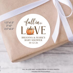 May include: A round white sticker featuring a watercolor orange pumpkin replacing the letter O in the text fall in LOVE. Below the pumpkin, the text reads BRIANNA & MARK'S BABY SHOWER 10-12-26. Available sizes listed are 1.5"x1.5", 2"x2", and 2.5"x2.5".