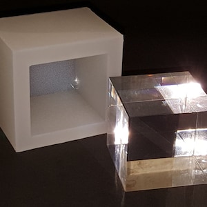Può includere: Un cubo di vetro trasparente si trova all'interno di uno stampo in silicone bianco. Il cubo riflette la luce, creando un effetto arcobaleno.