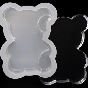 May include: A translucent, flexible silicone mold shaped like a teddy bear, paired with a clear, rigid acrylic lid of the same bear silhouette. The mold features a hollow interior for casting, while the lid provides a flat, matching cover.