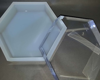 Pode incluir: Uma forma hexagonal de silicone com uma tampa de plástico transparente. A forma é cinza claro e a tampa é transparente.
