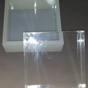 May include: A clear acrylic box with a gray silicone mold inside. The mold is square and has a smooth surface.