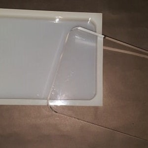 Puede incluir: Un molde de silicona blanco con una tapa de acrílico transparente. El molde es rectangular y tiene una superficie lisa. La tapa es ligeramente más grande que el molde y tiene un borde biselado.