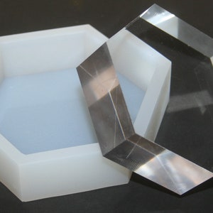 Puede incluir: Un molde hexagonal de silicona blanco con una tapa hexagonal de acrílico transparente. El molde está abierto y la tapa está a un lado.