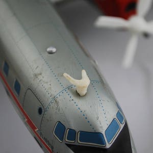 以下が含まれることがあります： 青い窓と赤いストライプのある銀色の飛行機のおもちゃ。飛行機の上には白いプラスチックのプロペラが付いています。