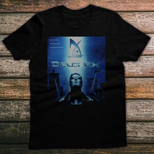 May include: Black t-shirt featuring a blue graphic from the video game Deus Ex. The design includes the game's logo, the title "Deus Ex", and a stylized image of a person in front of a cityscape.