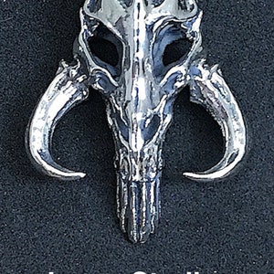 Puede incluir: Un gran colgante de plata de ley en forma de casco mandaloriano. El colgante está detallado con un diseño de calavera y dos cuernos.