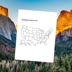 May include: A white sheet of paper featuring the text "Reading through the USA" and a black outline map of the United States. The background shows a mountain range and a forest.