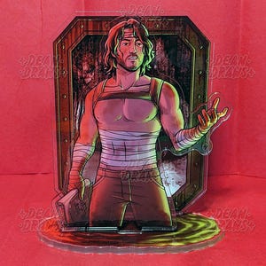 May include: A clear acrylic figure of a man with long hair, wearing a tank top, bandages, and brown pants. The figure is set against a background of a wooden door and a red surface. The base of the figure is a green and yellow liquid.