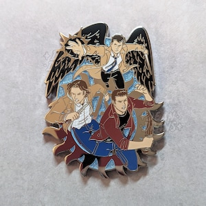 May include: Enamel pin depicting three figures in action poses, with a character in a tan trench coat and black wings. The pin has a blue and gold colour scheme with glitter accents.