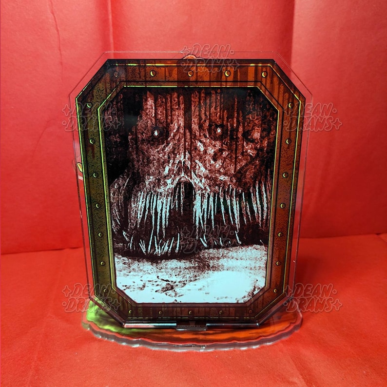 May include: A clear acrylic display featuring a horror-themed illustration. The artwork depicts a monstrous face with sharp teeth and glowing eyes within a wooden frame. The base is a translucent green, set against a red background.