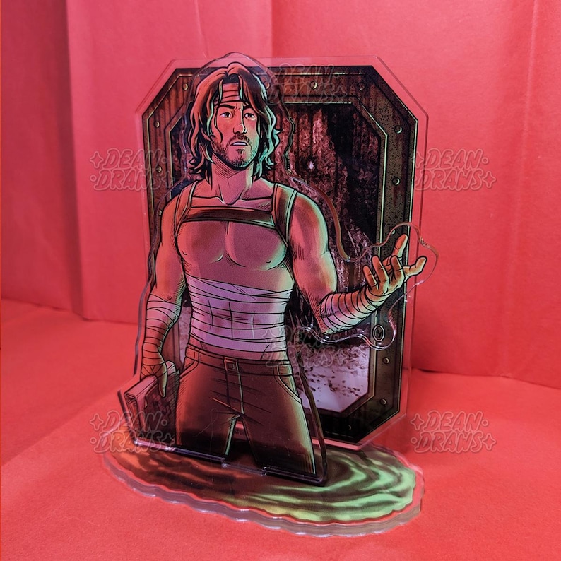 May include: A clear acrylic figure of a man with long hair, a headband, and wrapped bandages. He wears a harness and pants, holding a book. The background features a door and a green, glowing liquid base. The figure is a stylized illustration.