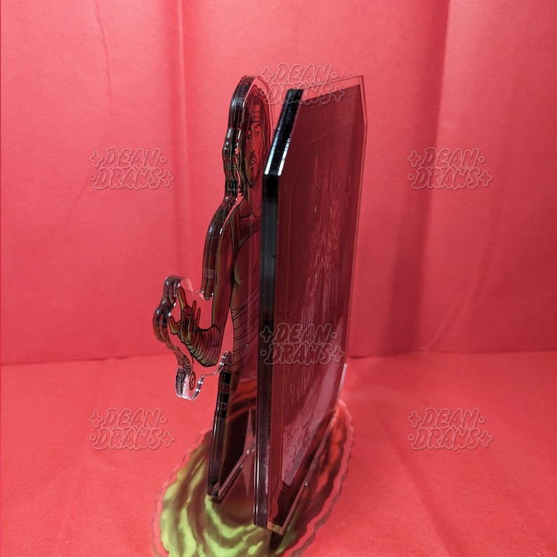 May include: A clear acrylic stand-up figure featuring a character with a head wrap and arm wraps. The figure is standing in front of a black rectangular panel. The base has green and yellow accents. The words "DEAN DRAWS" are printed on the stand.