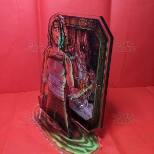 May include: A clear acrylic figure featuring a male character with long hair, bandages, and a sleeveless top. The figure is standing in front of a wooden door with a forest scene visible. The base is a green and yellow swirl.