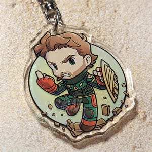 May include: A clear acrylic keychain featuring a cartoon illustration of a character in a green and orange outfit, holding a shield. The character has brown hair and a beard, with a determined expression. The keychain is attached to a silver chain.
