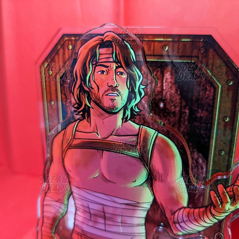 May include: A clear acrylic figure featuring a male character with long brown hair, a headband, and wrapped bandages. The figure has a muscular build and is set against a red background with a green and brown border.