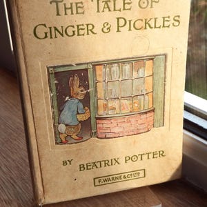 May include: A vintage book titled "The Tale of Ginger & Pickles" by Beatrix Potter. The cover features a brown rabbit in a blue coat entering a shop with a brick facade and a window filled with jars.