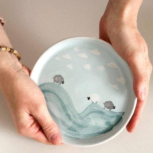Puede incluir: Un plato redondo de cerámica con fondo azul claro y diseño caprichoso. El plato presenta tres ovejas sobre una colina verde estilizada con nubes blancas. El plato está sostenido por dos manos.