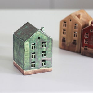 May include: Three miniature ceramic houses with different colored glazes. The houses are all the same size and shape, but have different colored roofs and walls. The houses are all sitting on a white surface.