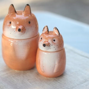 Peut inclure: Deux figurines de renard en céramique, l'une plus grande que l'autre, avec un glaçage orange et blanc. Les renards ont des yeux et des nez noirs.