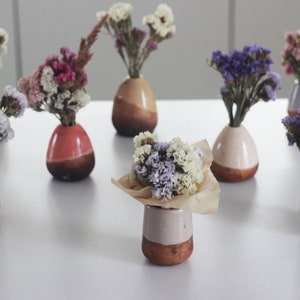 Op de afbeelding: Een verzameling kleine keramische vazen in verschillende kleuren, elk gevuld met gedroogde bloemen. De vazen zijn allemaal in verschillende vormen en maten, en de bloemen zijn een mix van paars, wit en roze.