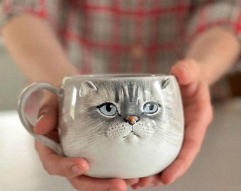May include: A ceramic mug featuring a sculpted, three-dimensional cat face with grey and white fur, light blue eyes, and a small pink nose. The mug has a rounded body and a curved handle on the left side, showcasing detailed painted whiskers.