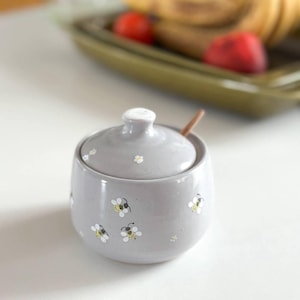 Puede incluir: Un azucarero de cerámica gris claro con tapa a juego y una pequeña cuchara de madera. El exterior del cuenco y la tapa están decorados con un patrón repetitivo de pequeñas abejas pintadas a mano en amarillo y negro y flores blancas.