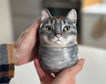 May include: A ceramic jar shaped like a grey tabby cat with a rounded body, pointed ears, and detailed facial features including large green eyes, a pink nose, and white whiskers painted against a textured grey and white striped fur pattern.