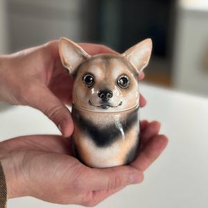 Pode incluir: Um pote de cerâmica em forma de cabeça de cão com orelhas grandes e pontiagudas. O cão tem o rosto castanho-claro, olhos castanhos escuros, nariz preto e uma zona do pescoço preta e castanha. O pote apresenta uma costura horizontal central.