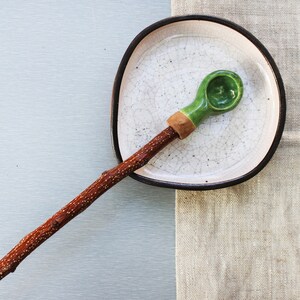 May include: A green ceramic spoon with a wooden handle resting on a white ceramic plate with a black rim. The plate is on a light brown linen surface.