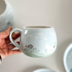 Puede incluir: Una taza de cerámica azul claro con forma redondeada y asa. La taza presenta una escena pastoral con una oveja, nubes y hierba verde. Se ven platos a juego adicionales.