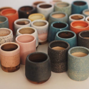 Op de afbeelding: Een verzameling kleine, kleurrijke keramische potten in verschillende tinten blauw, roze, grijs en wit. De potten zijn gevlekt en hebben een rustieke, handgemaakte uitstraling.