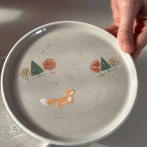 May include: A round ceramic plate with a light gray background featuring a painted orange fox walking along a dotted path. The scene includes small green pine trees and orange autumn trees with scattered leaves falling across the surface.