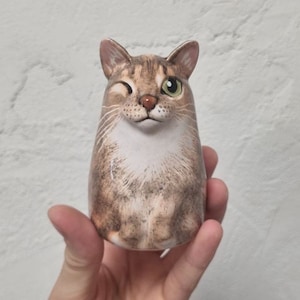 以下が含まれることがあります： 白の模様のある茶トラ猫のセラミック製置物。猫は片目を閉じ、いたずらっぽい表情をしています。